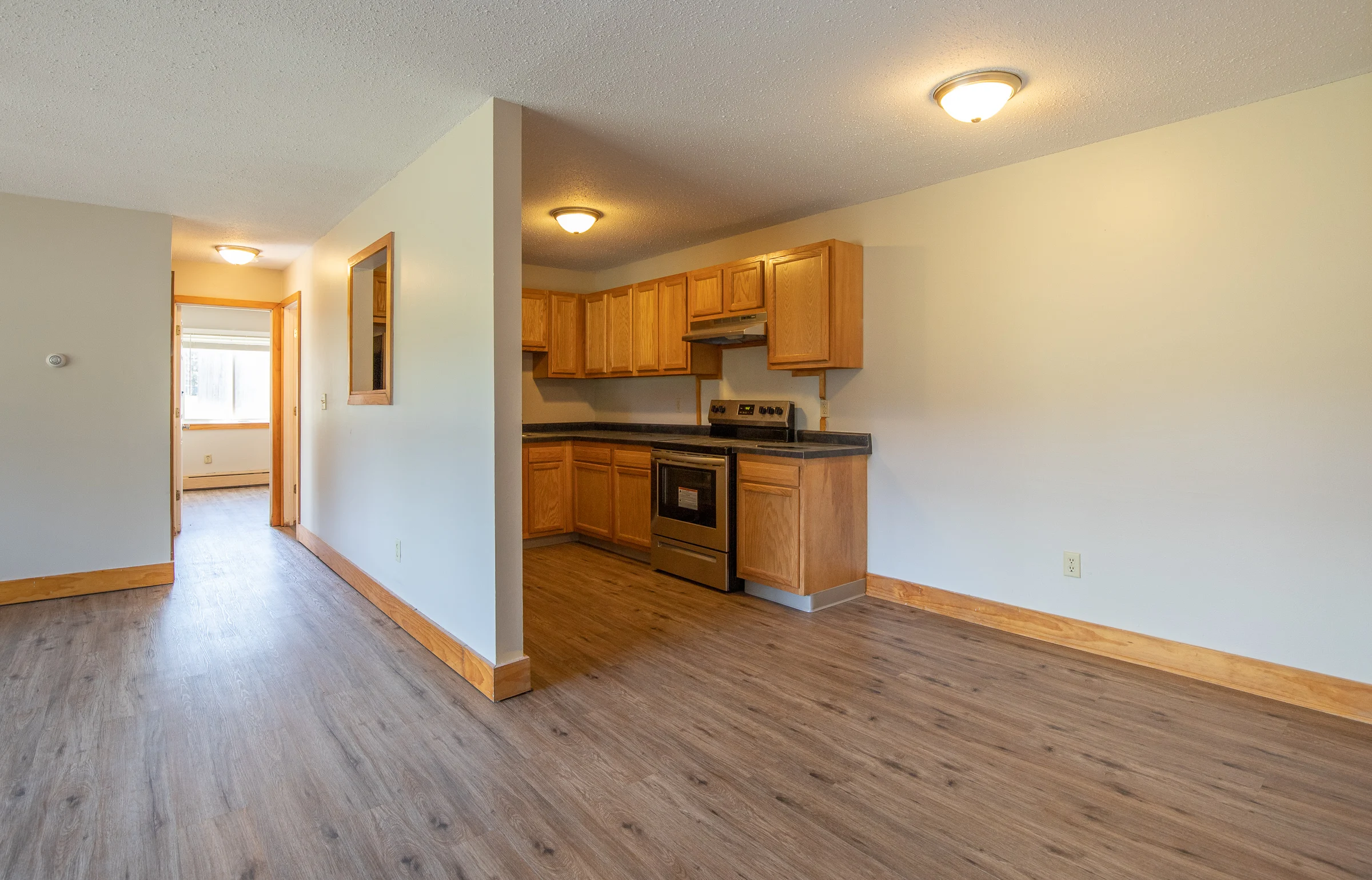 An open kitchen and living area with wooden cabinets and a modern stove. The flooring is a light brown laminate. Natural light enters through a window on the left, leading to a hallway.