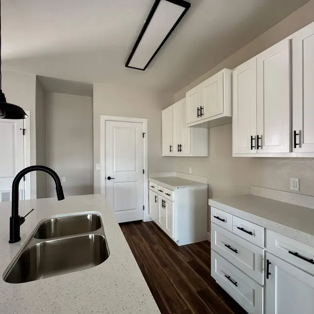 A modern kitchen featuring white cabinetry, a large island with a double sink, and a sleek black pendant light. The countertops are light-colored, complementing the overall bright aesthetic. The space has wooden flooring and a door leading to another area.