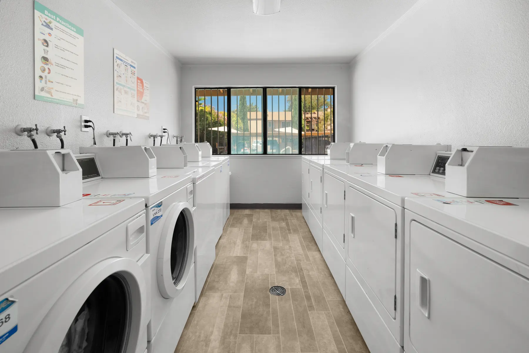 A clean and well-lit laundry room featuring multiple washing machines and dryers arranged neatly in rows. Large windows allow natural light to flood the space, illuminating the light-colored walls and flooring.