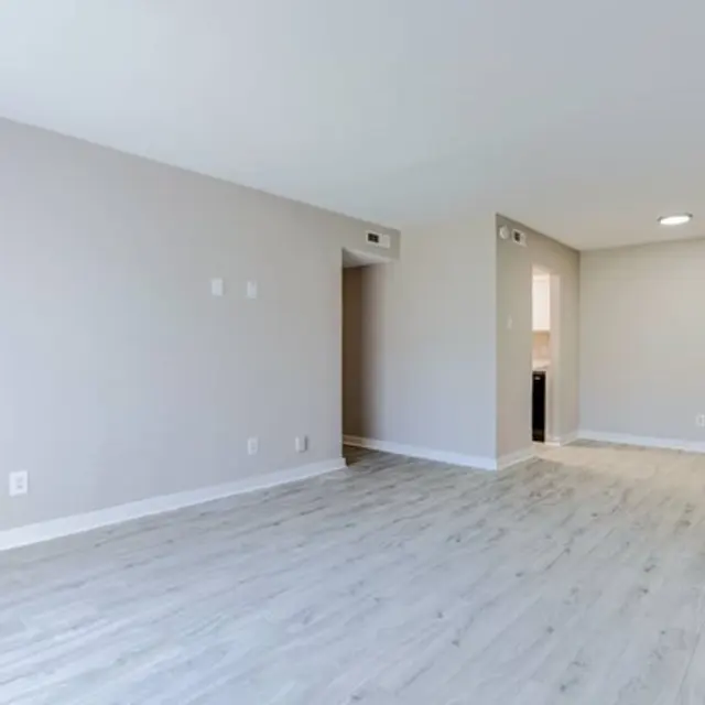 An empty apartment living room with light-colored wood floors and neutral walls. There is a doorway leading to a kitchen on the right, and a small hallway on the left.