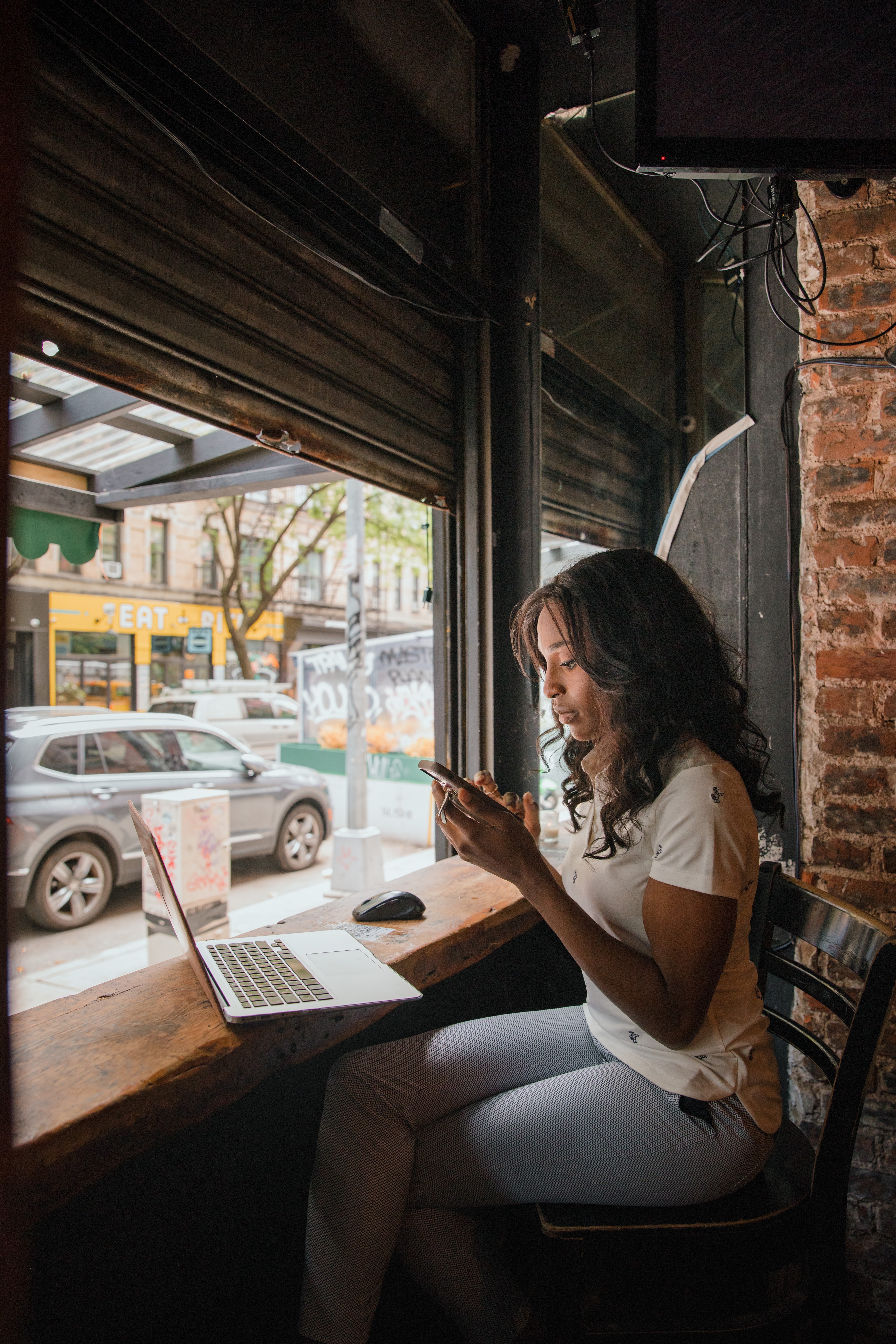 Café Workspace A woman with long dark hair sits by a large window in a café, holding a smartphone and looking at it. A laptop is open on the table in front of her, with a view of the street visible outside.
