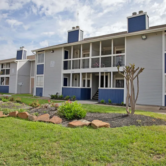 Exterior view of a two-story apartment complex with blue and gray siding, surrounded by landscaped grounds and a parking area.