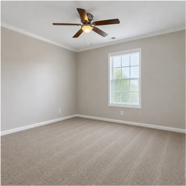 An empty room featuring light beige walls and a tan carpet, equipped with a ceiling fan and a window that lets in natural light.