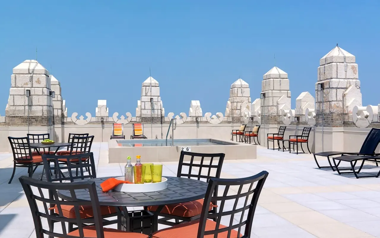 A rooftop terrace featuring stylish black metal chairs with orange cushions around a circular table. There are drinks on the table, and in the background, ornate architectural elements with a blue sky above.