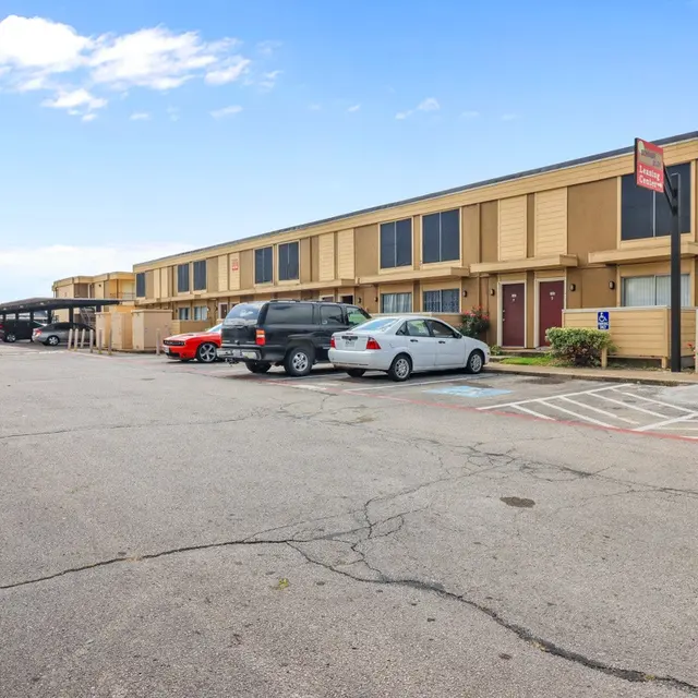 View of a low-rise apartment complex with parking lot, featuring several vehicles and blue sky in the background.