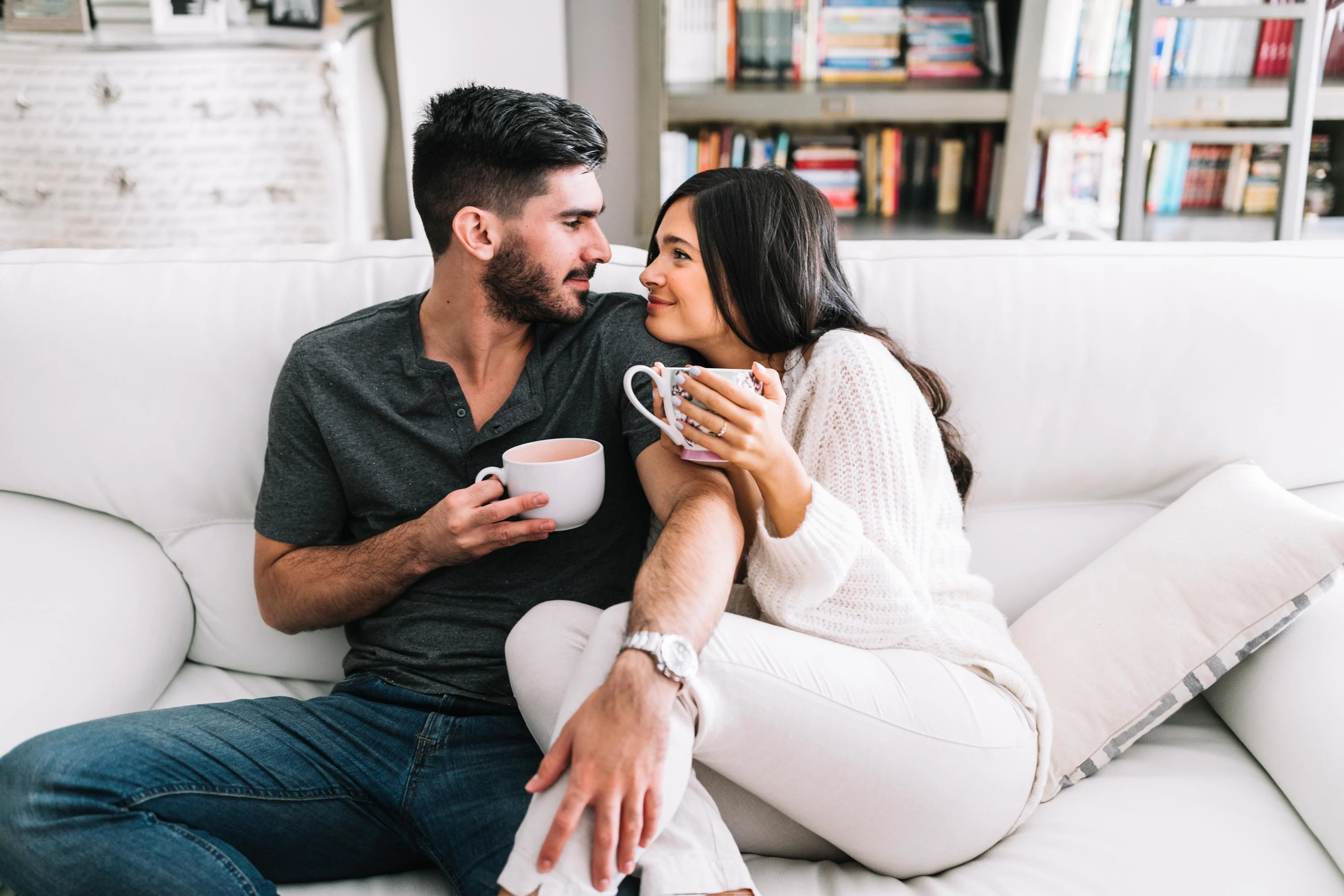 Cozy Couple Moment A young couple sitting on a white couch, holding cups of coffee, gazing at each other with smiles. The setting is cozy with bookshelves in the background.