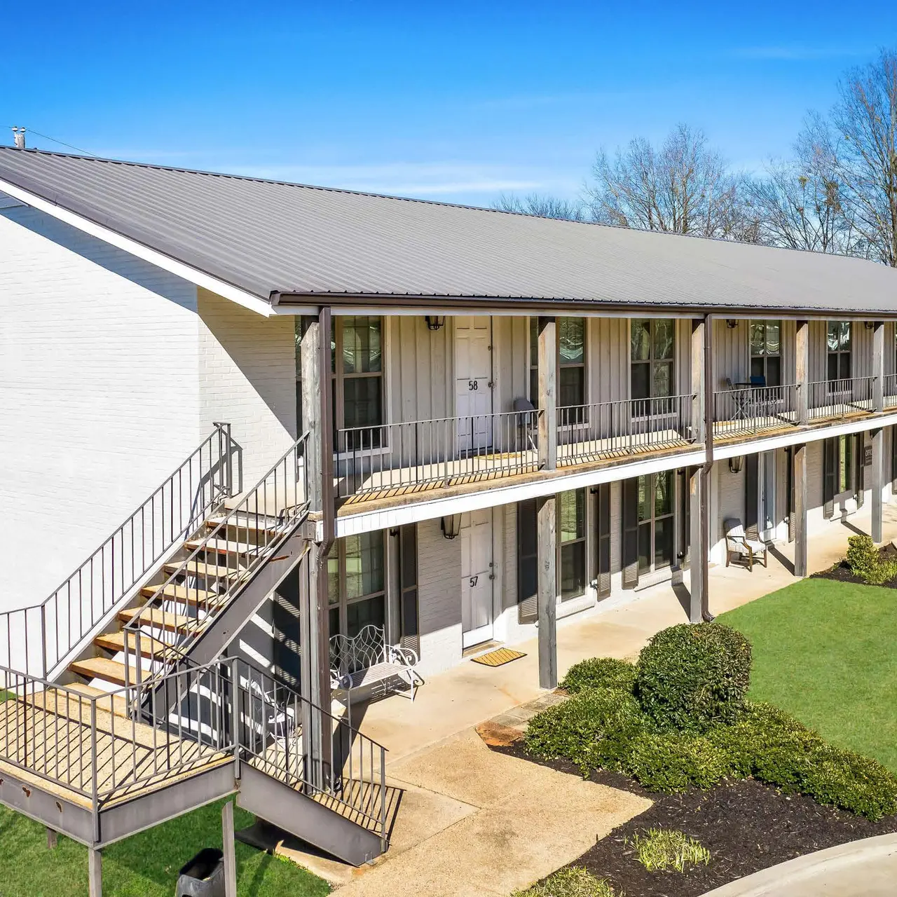 A two-story apartment building with a metal roof, featuring a wooden staircase leading to the upper level. Ground level has open doors and windows. Green lawn surrounds the building with some trees in the background.