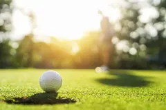 Golf Ball in Evening Light A golf ball sits on lush green grass with a blurred figure of a golfer in the background, illuminated by warm golden sunlight.
