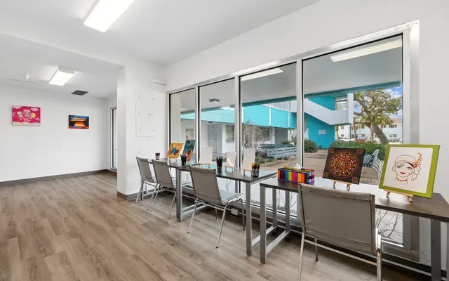 A bright and welcoming art space with a long table adorned with art supplies and paintings. Large windows offer natural light and a view of the outdoors. The interior has wooden flooring and features various artworks on the walls.