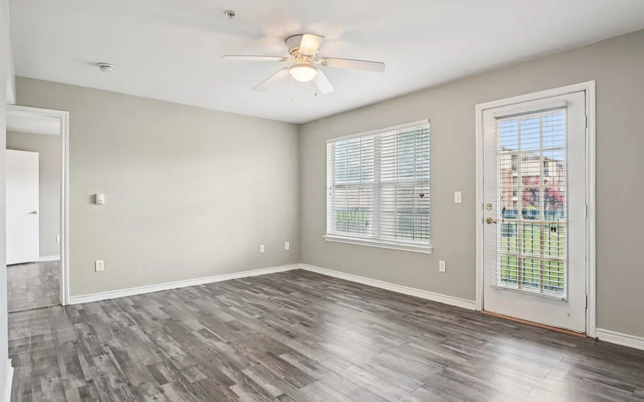 An empty room with a ceiling fan, light-colored walls, and gray wooden flooring. There is a window with white blinds, and a door leading outside. The room is well-lit and spacious.