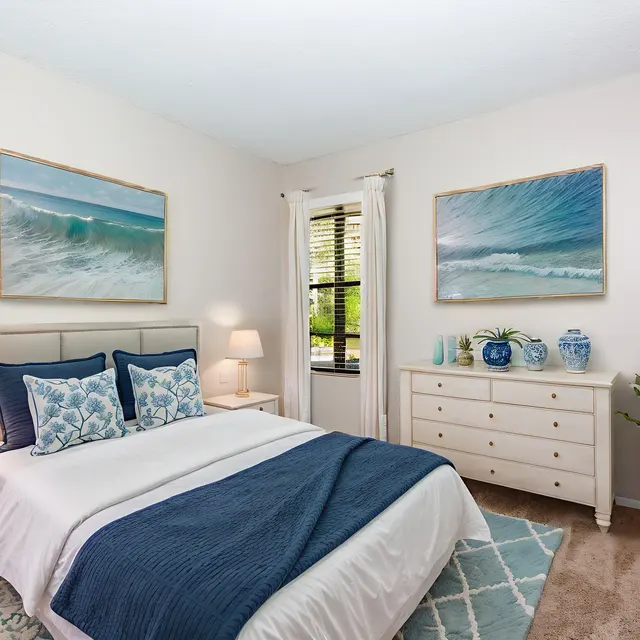 A beautifully decorated bedroom featuring a bed with blue and white linens, two wall paintings of ocean waves, a bedside table with a lamp, and a dresser adorned with vases and a potted plant.