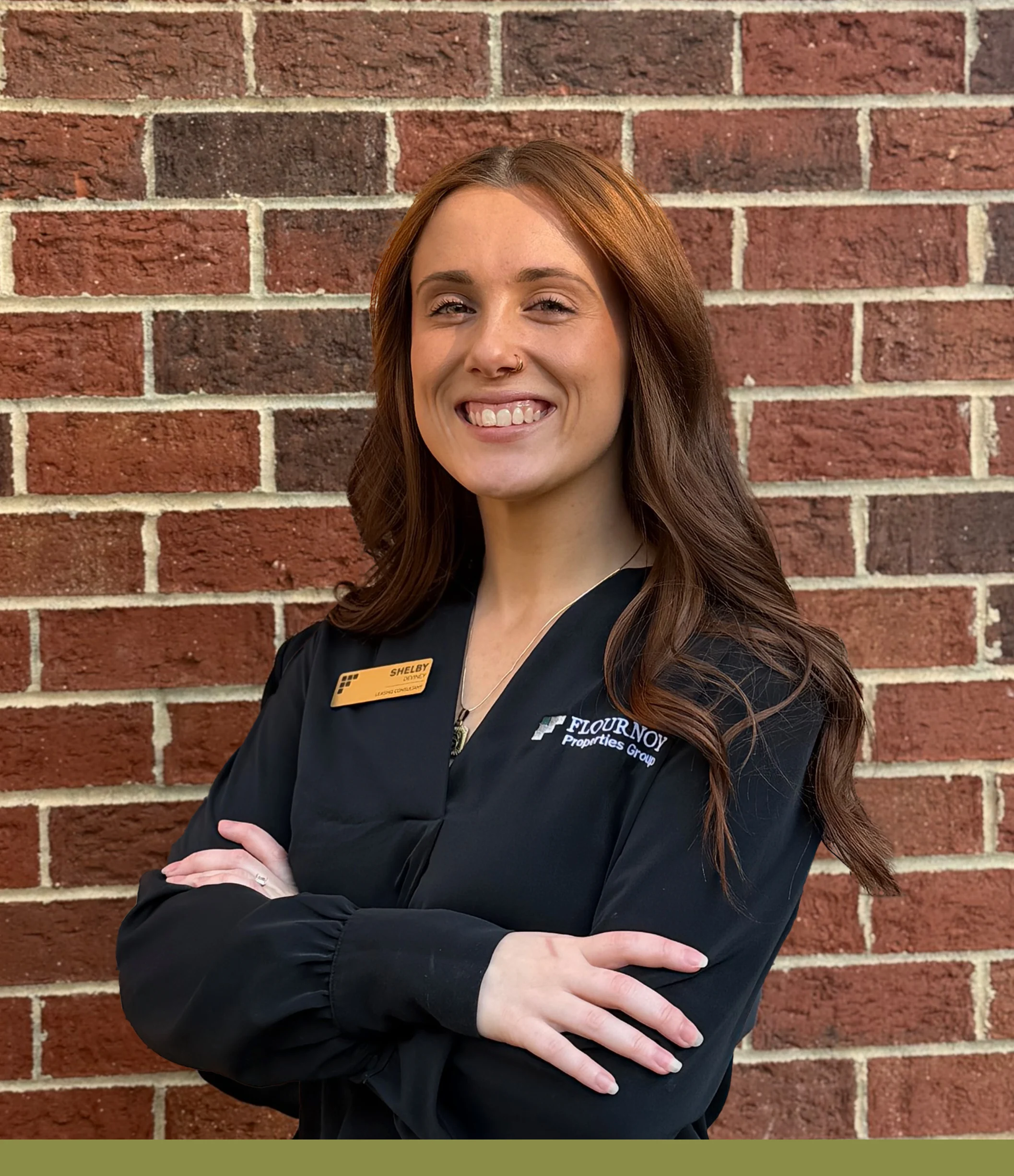 Confident Professional Portrait A woman smiling confidently with her arms crossed, wearing a black professional outfit, against a brick wall background.