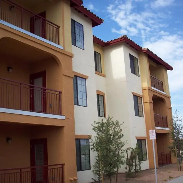 Colorful Apartment Building A multi-story apartment building with a combination of yellow and cream-colored walls, featuring balconies and red railings. There are trees and shrubs in front, alongside a street sign.