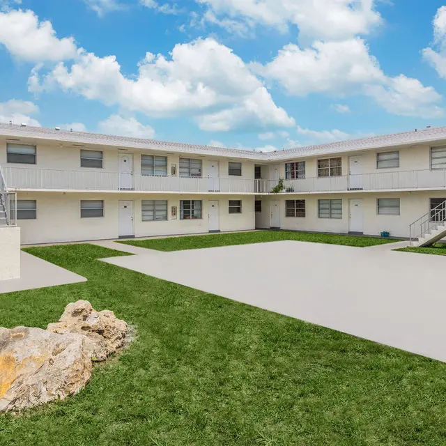 Exterior view of a modern multi-unit apartment complex featuring a landscaped courtyard and white buildings against a blue sky with clouds.