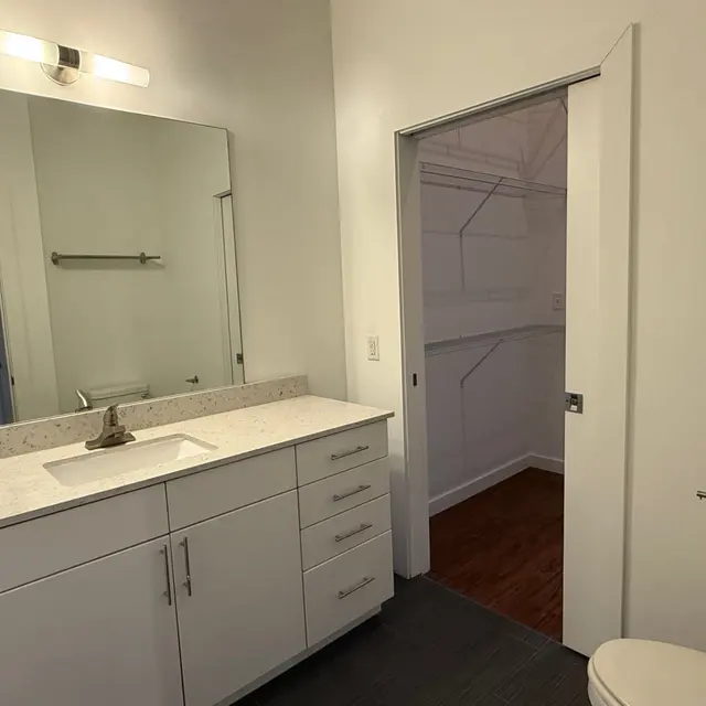 A modern bathroom featuring a white vanity with a sink, a large mirror, and a doorway leading to a closet. The bathroom is well-lit, and the closet is partially visible.