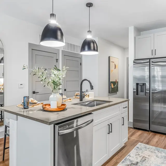 A contemporary kitchen featuring gray cabinets, a stainless steel refrigerator, and pendant lighting. There's a sink with a modern faucet and a small table with dining chairs visible in the background.