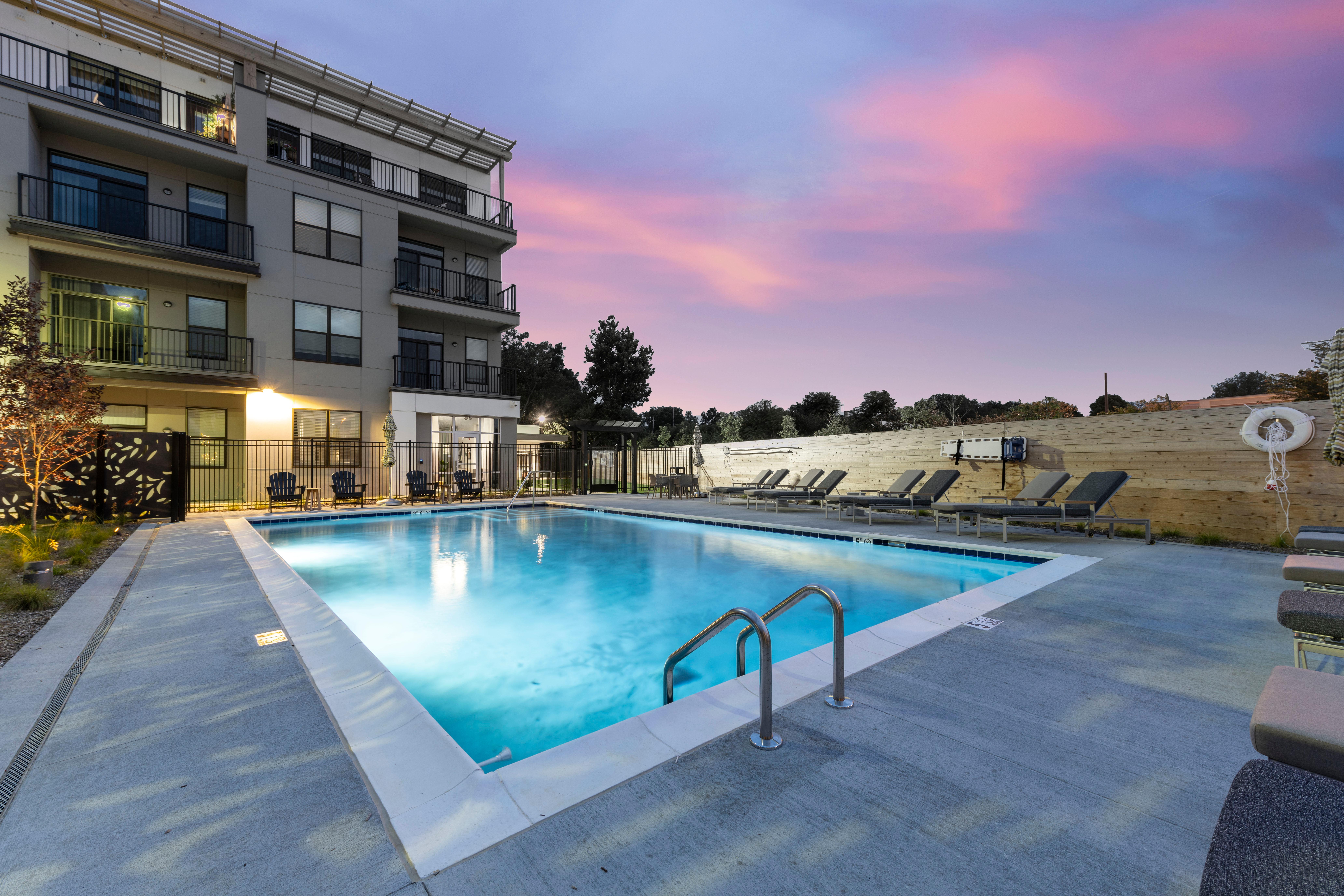 View of a modern swimming pool area at dusk, surrounded by a multi-story apartment building and lounge chairs.