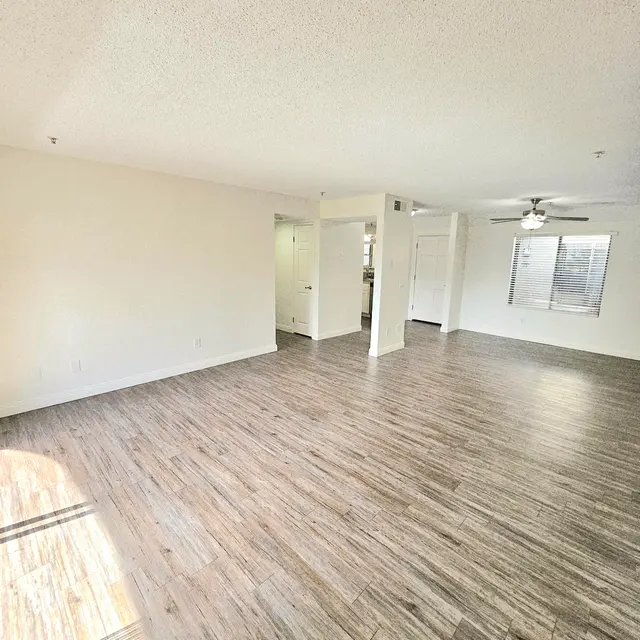 A spacious living room with light-colored walls and wooden flooring, featuring a ceiling fan and a large window, providing ample natural light.