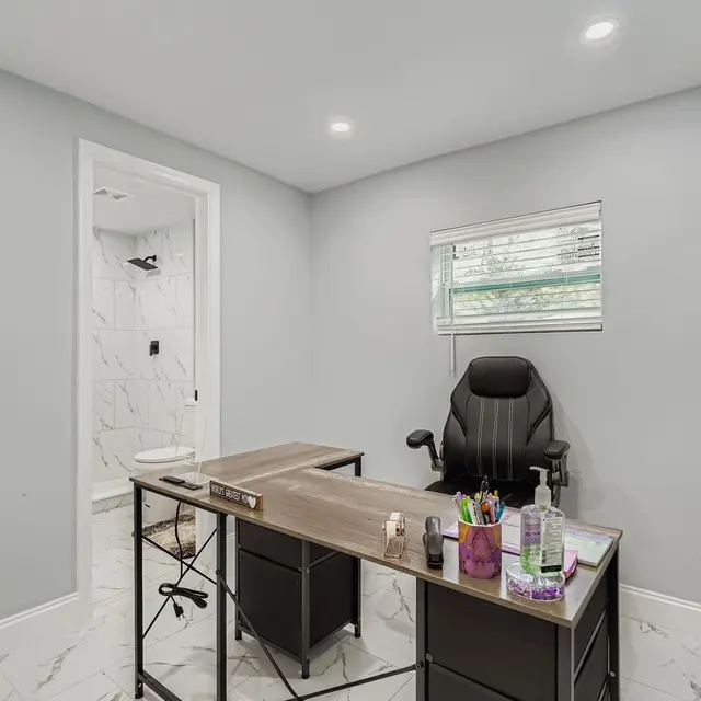 A modern home office featuring a minimalistic desk and an ergonomic chair. The walls are painted light gray and the floor is tiled with a marble pattern. A window provides natural light. A door leads to a bathroom area.