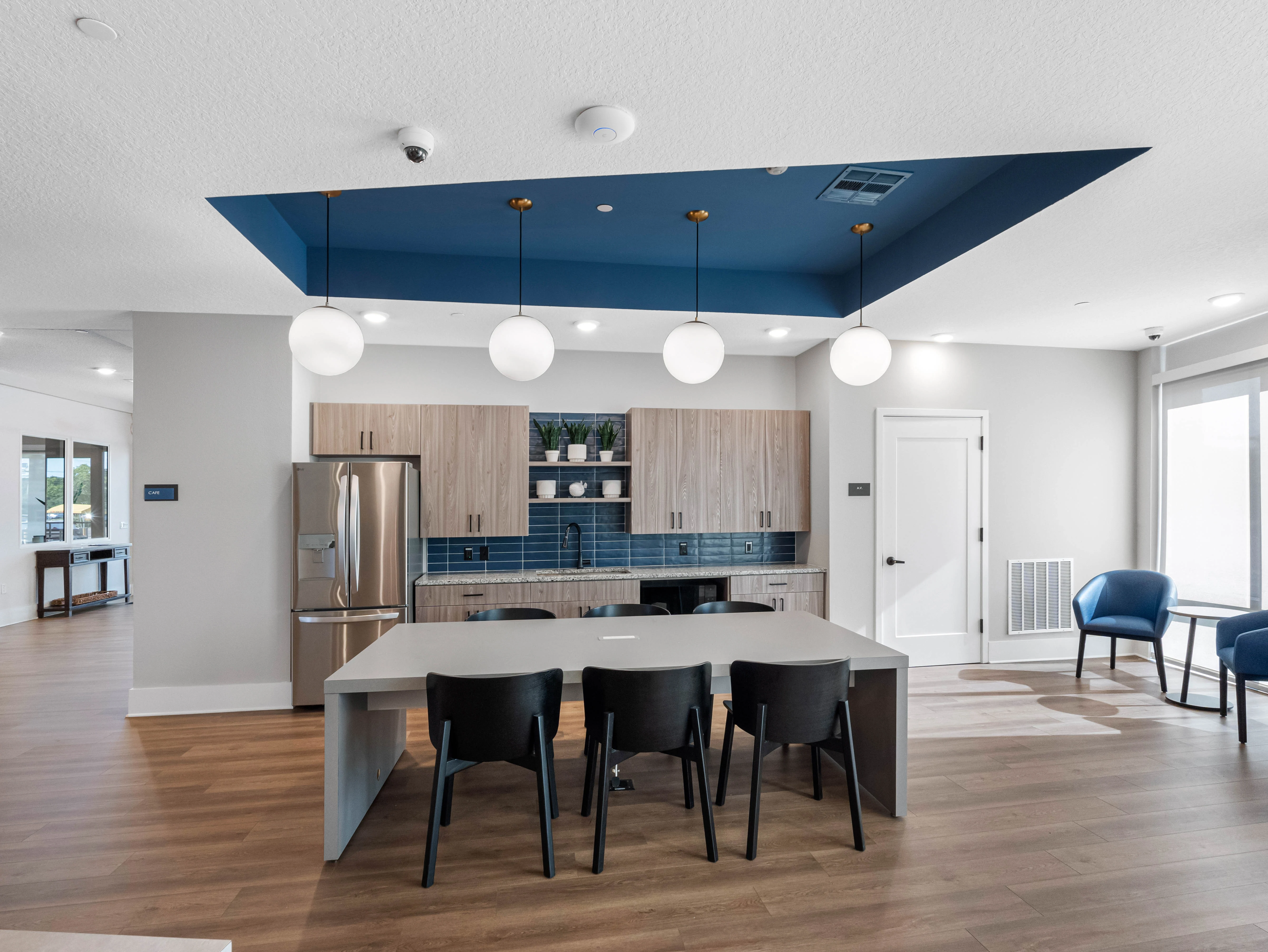 A modern kitchen area featuring a large island with black chairs, pendant lighting, and a fridge. The cabinets are light wood with blue tile backsplash.