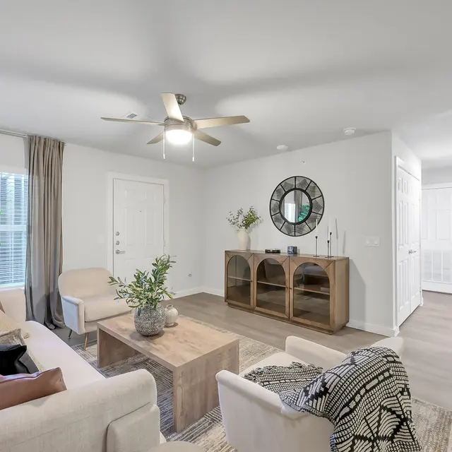A modern living room featuring a light-colored sofa, a wooden coffee table, and a stylish sideboard with decor. There are large windows with curtains, a decorative plant, and a round mirror on the wall. The flooring is light wood and the room has a ceiling fan.