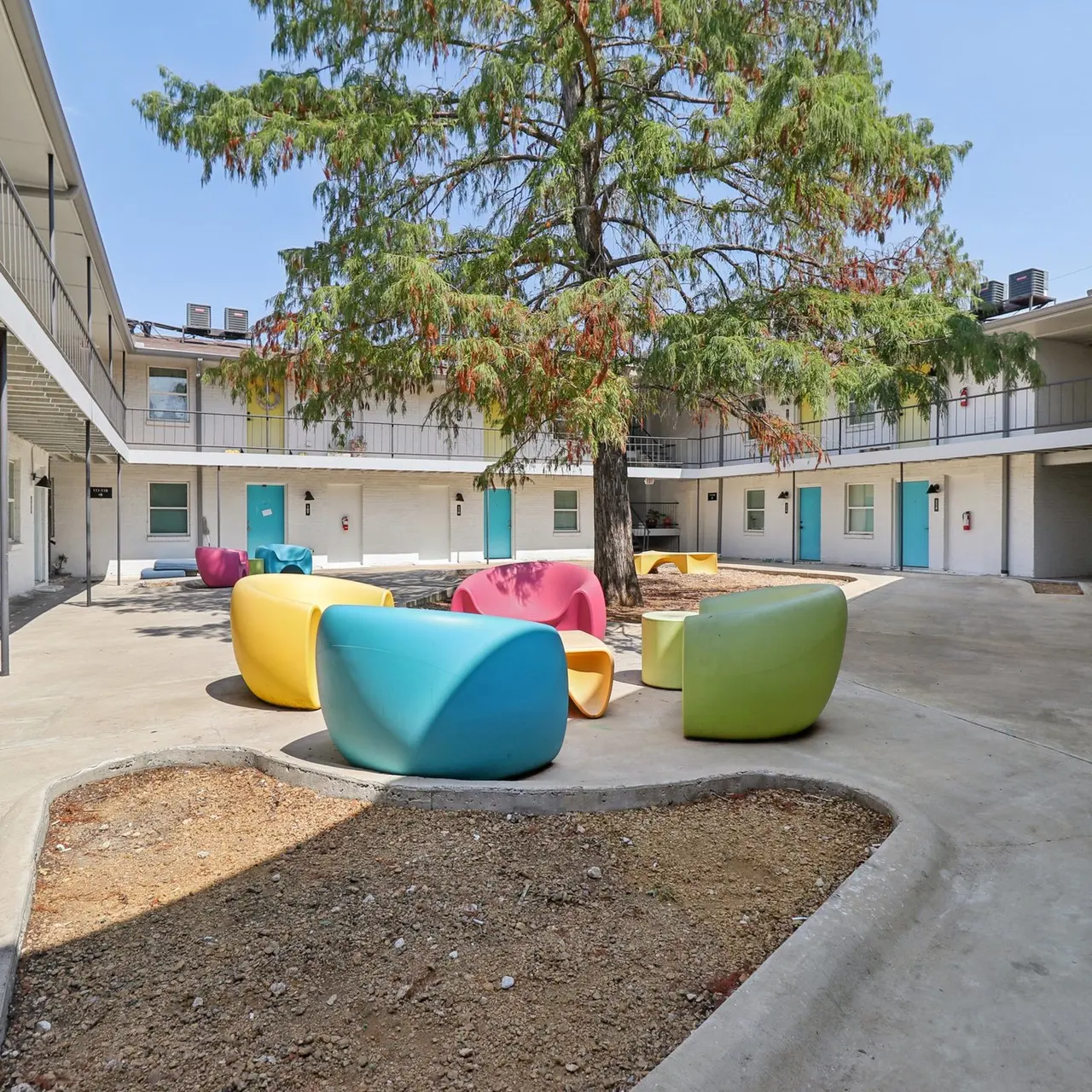 A colorful courtyard surrounded by building facades, featuring a large tree and vibrant seating areas in various shapes.