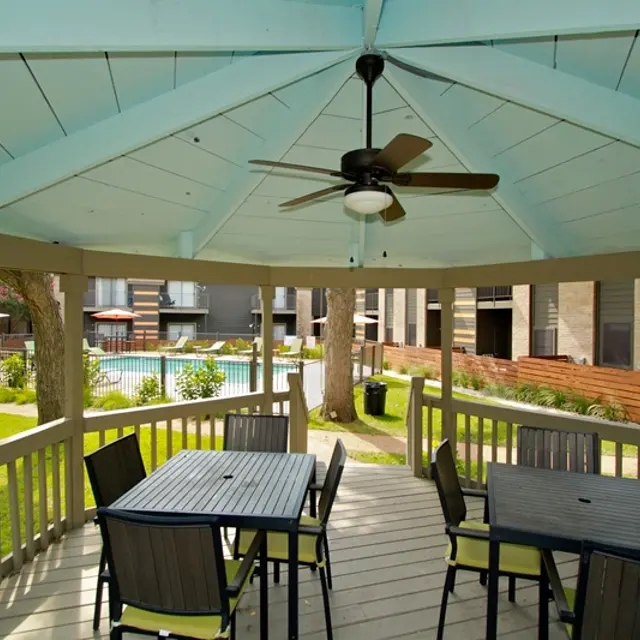 A spacious outdoor pavilion with a fan and tables, overlooking a swimming pool and greenery.