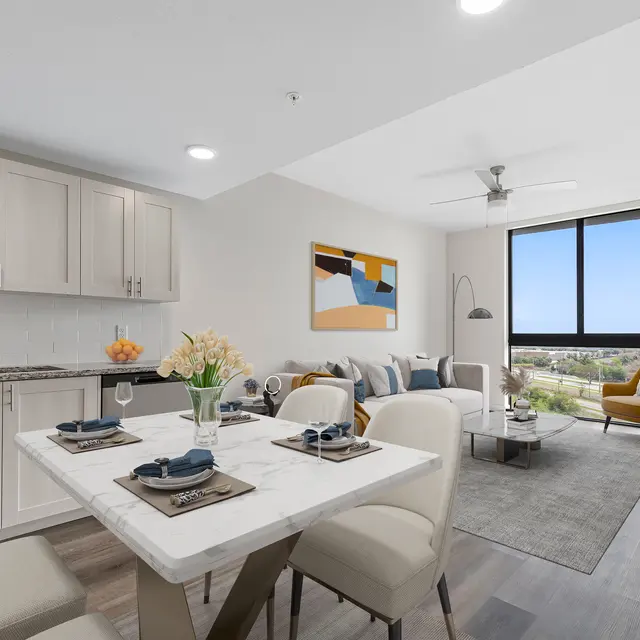A modern apartment interior featuring a kitchen and living area, with a dining table set for two and a cozy couch with decorative pillows. A large window offers a view of the outdoors.