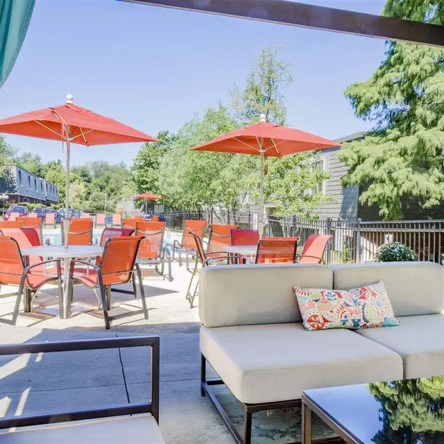 Charming Outdoor Lounge Area by the Pool An outdoor lounge area featuring a modern sofa with decorative pillows, looking out toward a pool with red umbrellas and tables.