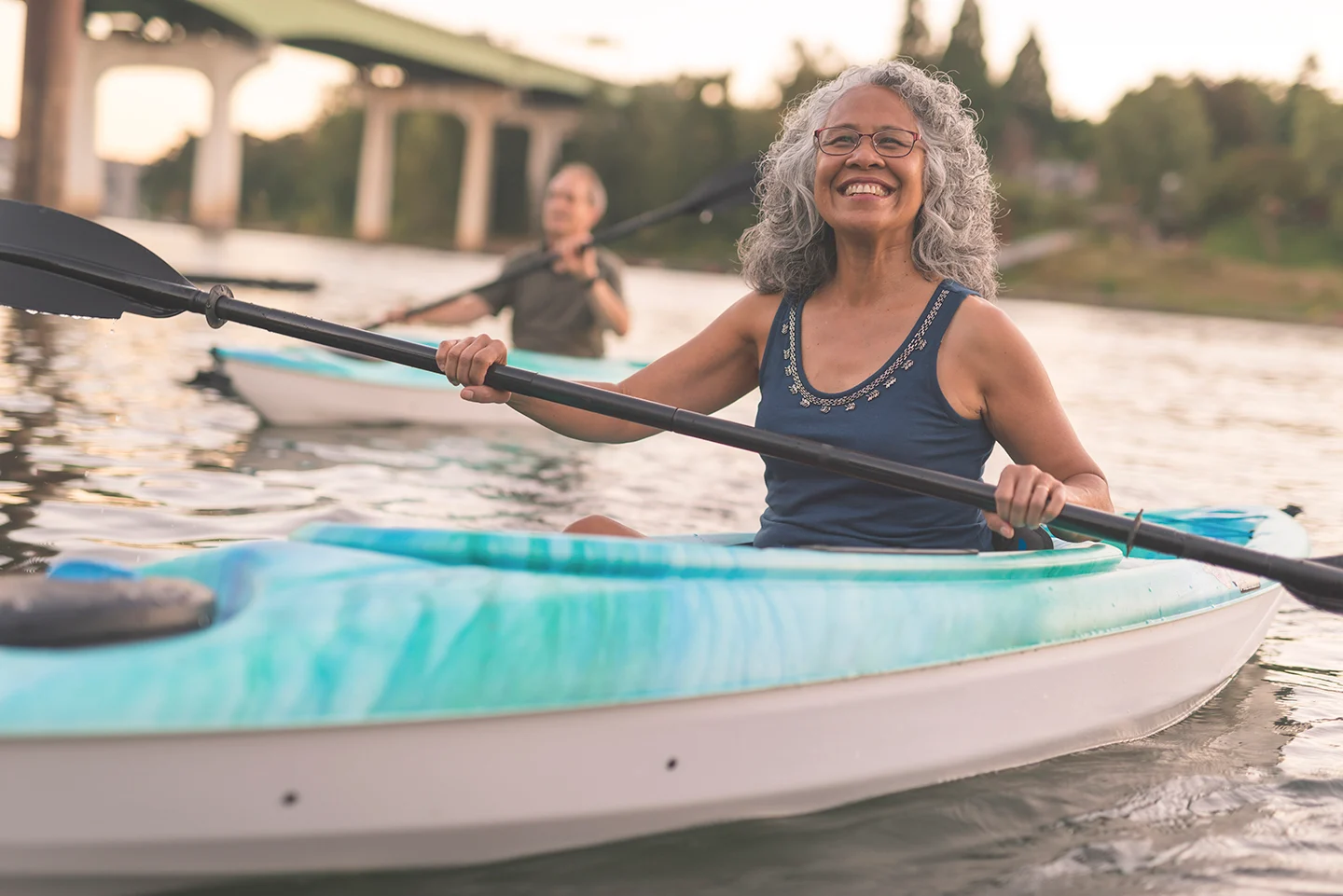 Kayaking Adventure A woman with curly gray hair smiles while kayaking on a calm waterway. In the background, another person in a kayak is visible. The setting features a bridge and greenery.