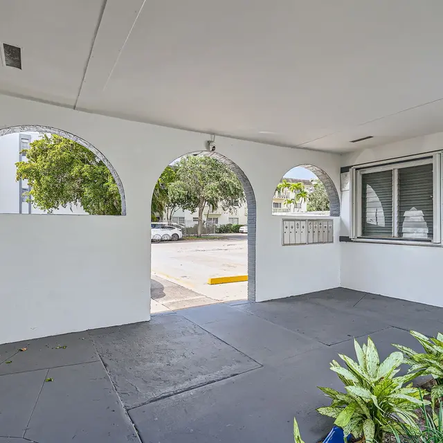 An outdoor entryway with a concrete floor, featuring arches that frame the view of a landscaped area and parking lot. The wall has a mailbox setup and a door leading inside. A small plant is visible in the foreground.