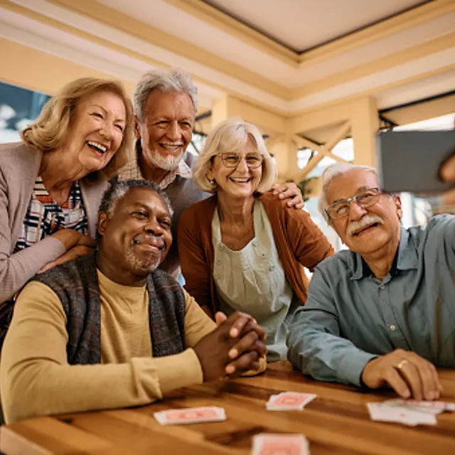 A group of five elderly friends taking a selfie together at a wooden table, smiling and having a good time.