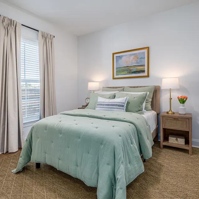 Cozy bedroom with a green quilted bedspread, two bedside lamps, a framed painting, and light curtains. Natural light from the window.