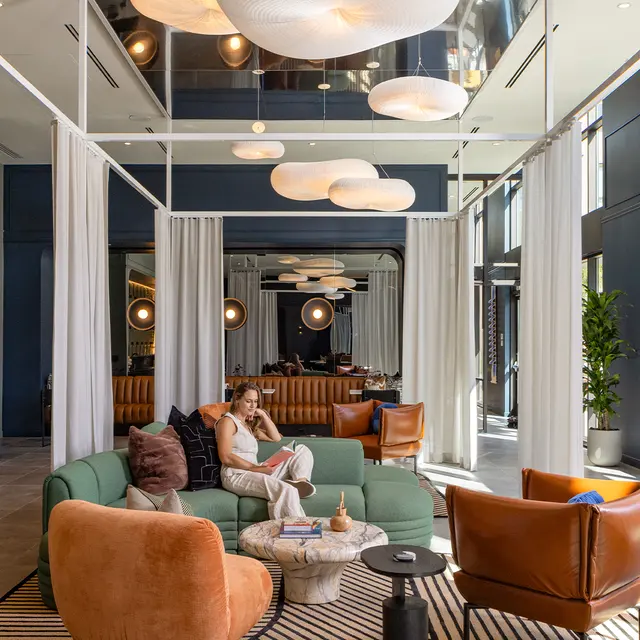 A stylish modern lobby with a mix of seating options, featuring a green sofa, brown armchairs, and a decorative coffee table. Light fixtures hang from the ceiling, and large windows allow natural light to fill the space.