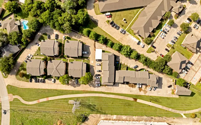Aerial view of an apartment complex with several buildings, parking areas, and green spaces. A swimming pool is visible to the left, and a road curves around the property.