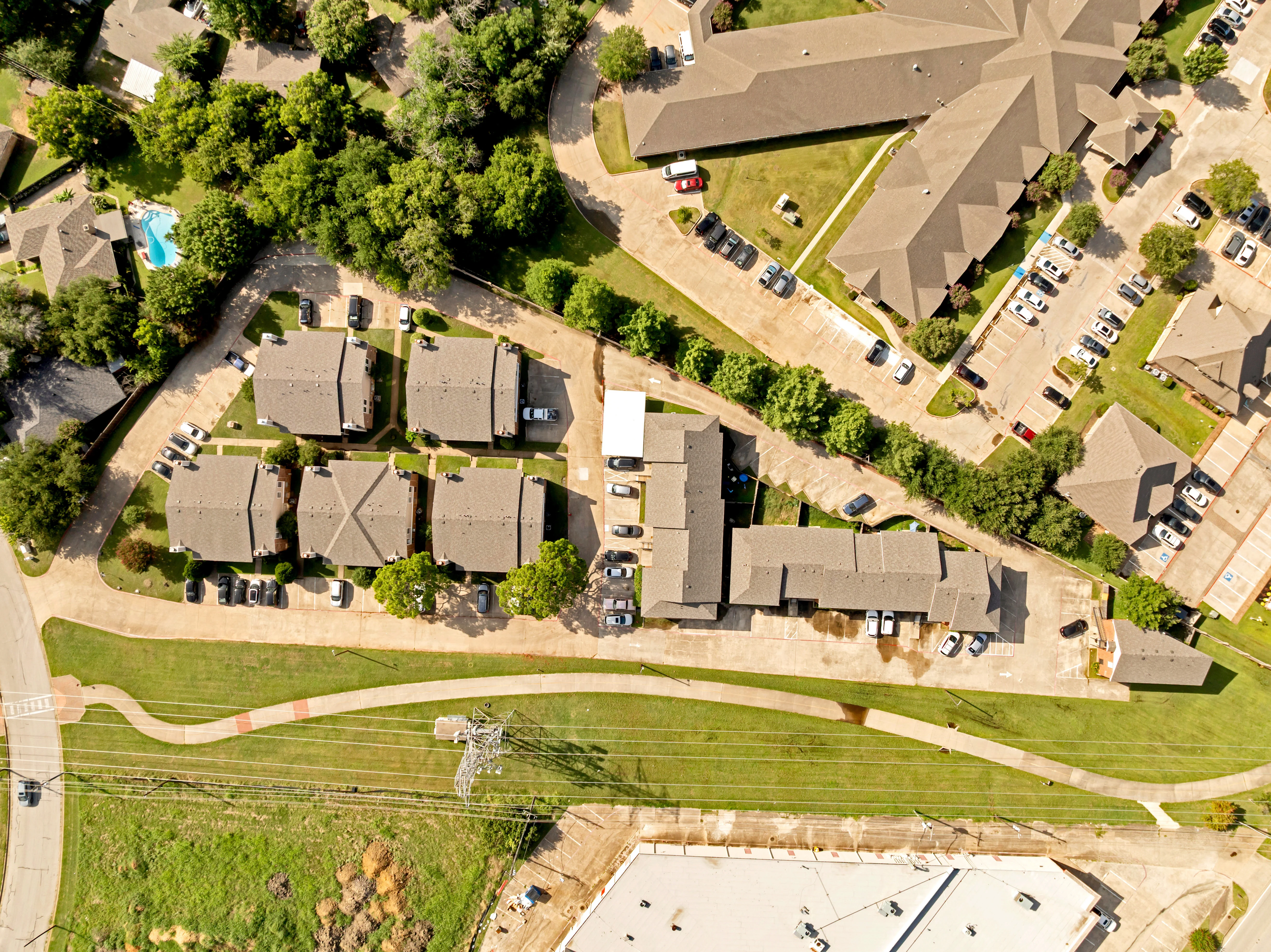 Aerial view of an apartment complex with several buildings, parking areas, and green spaces. A swimming pool is visible to the left, and a road curves around the property.