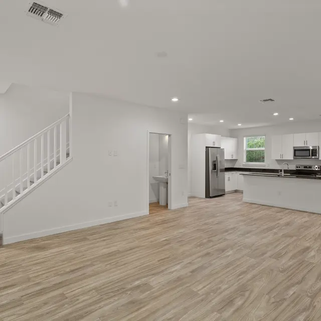 Interior view of a spacious, modern open-plan living area with wooden flooring, a staircase in the corner, and a kitchen with stainless steel appliances.