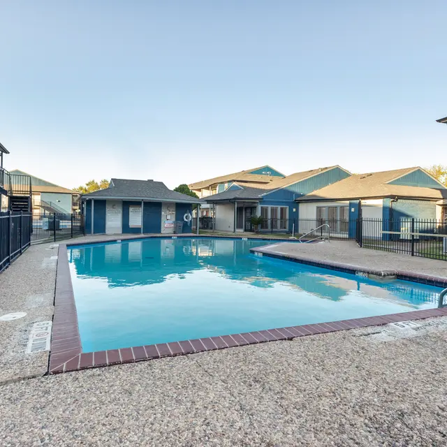 A clear swimming pool surrounded by textured concrete and fences, with apartment buildings in the background.