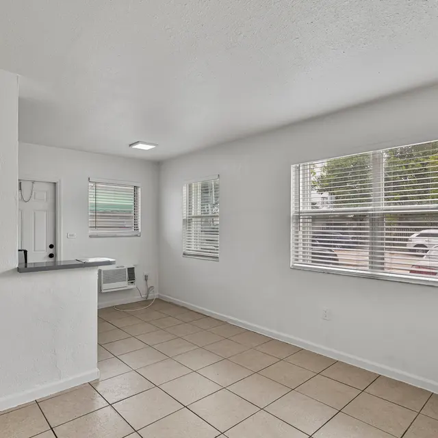 A clean and bright interior of a modern apartment with tiled flooring, white walls, and large windows allowing natural light to enter. A small kitchen area is visible to the left with a countertop.