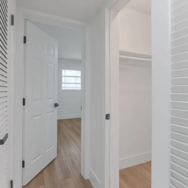A modern interior hallway with two open doors, leading to a room and a closet. The walls are painted white, and the floor has light wood planks. Natural light comes from a window in the room at the end of the hallway.