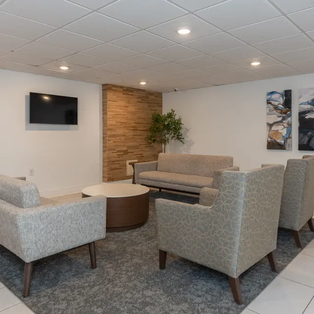 A modern lounge area featuring two light gray sofas, a round wooden coffee table, and two accent chairs. The walls include a textured wood panel and decorative artwork. A flat-screen TV is mounted on the wall.