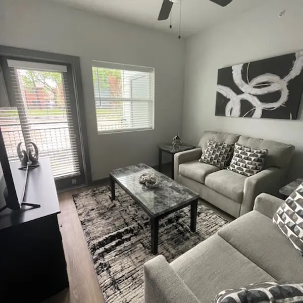A modern living room featuring a light gray sofa with patterned cushions, a black and white abstract wall art piece, a glass coffee table on a patterned rug, and a ceiling fan. There are windows allowing natural light in, and a dark wooden TV stand is visible.