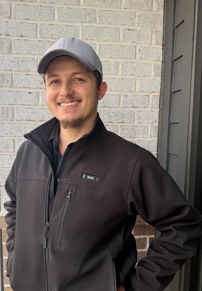 A young man wearing a gray cap and a black jacket stands outdoors, smiling at the camera with a brick wall in the background.