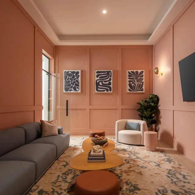 A modern living room featuring a pink wall, a blue sectional sofa, and decorative wall art. There is a round yellow coffee table, an orange ottoman, a small white armchair, and a green plant in a corner. A flat-screen TV is mounted on the wall opposite the sofa.