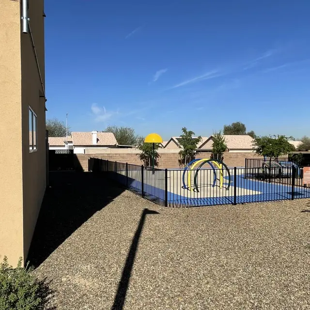 A playground enclosed by a black fence, featuring a colorful play structure, surrounded by trees and buildings under a clear blue sky.
