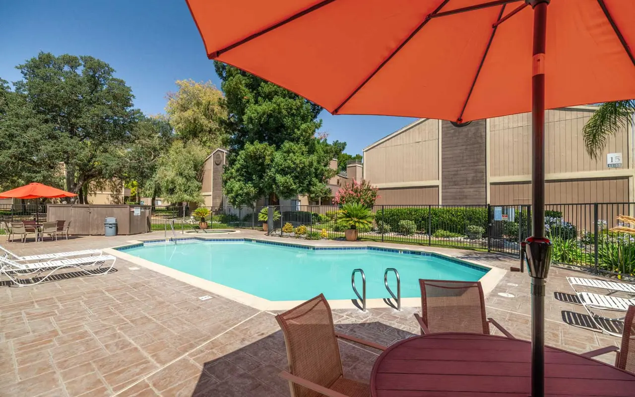 View of a swimming pool surrounded by lounge chairs and landscaped greenery, shaded by a large orange umbrella.