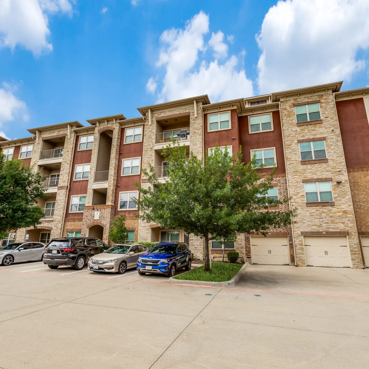 Exterior view of a modern apartment building with multiple levels, featuring brick and stucco architecture, trees, and cars parked in front.