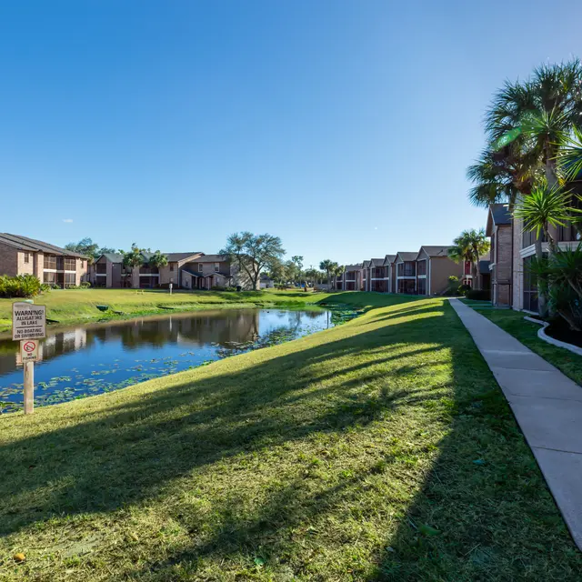 A peaceful view of an apartment complex along a waterway. There are palm trees and grassy areas lining a paved path next to the water.