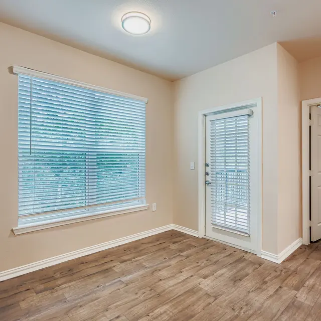 Empty Room with Wooden Flooring A light-colored empty room with wooden flooring, a large window with vertical blinds, and a door leading outside. The walls are painted in a neutral tone.