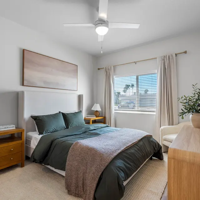 A cozy, modern bedroom featuring a large bed with green bedding, a decorative throw blanket, two bedside tables, a chair, and a window with drapes.